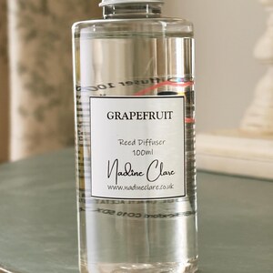 May include: A clear glass bottle with a silver cap, labelled "Grapefruit Reed Diffuser 100ml" by Nadine Clare. The bottle contains a clear liquid and is displayed on a light green surface. The website nadineclare.co.uk is also visible.
