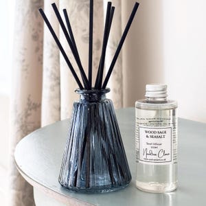 May include: A blue glass reed diffuser with black reeds and a clear 100ml bottle of Wood Sage & Seasalt fragrance. The bottle has a white label with text. The diffuser is on a light blue surface.