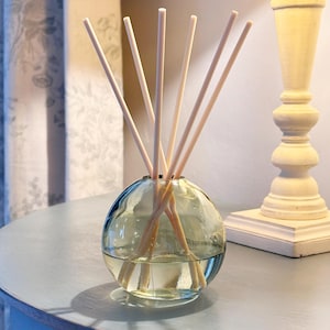 May include: A spherical glass diffuser with wooden reeds, filled with a clear liquid. The bottle is on a light blue surface, with a decorative lamp in the background. The reeds are designed to release fragrance.