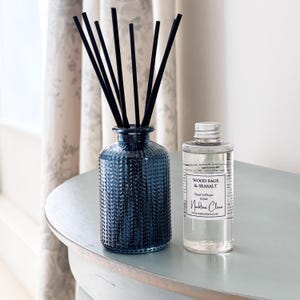 May include: A dark blue glass reed diffuser with black reeds and a clear bottle of Wood Sage & Seasalt reed diffuser liquid. The diffuser and liquid are on a light blue table.