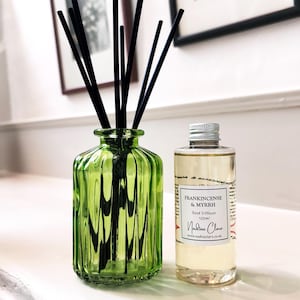 May include: A green glass reed diffuser with black reeds and a bottle of Frankincense & Myrrh Reed Diffuser. The bottle has a silver cap and a label with the text "Nadine Clare".