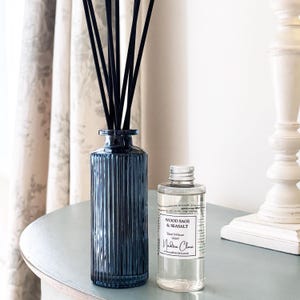 May include: A dark blue glass reed diffuser with black reeds and a bottle of Wood Sage & Seasalt reed diffuser oil. The diffuser has vertical lines and sits on a light blue table. The oil bottle has a white label.
