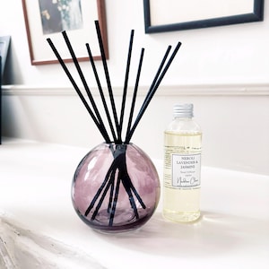 May include: A round, translucent purple glass reed diffuser vessel containing several thin black reeds, accompanied by a clear plastic bottle filled with light yellow liquid, labelled Neroli Lavender & Jasmine Reed Diffuser 100ml Nadine Claire.