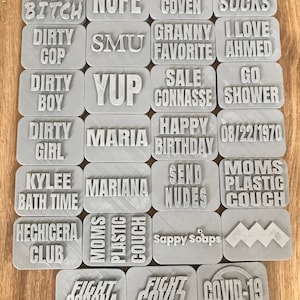 May include: A collection of gray rectangular objects with various words and phrases in raised lettering. Phrases include "Dirty Bitch", "Nope", "Hermosa Coven", "Covid Sucks", "Fight Corona", and "Covid-19".