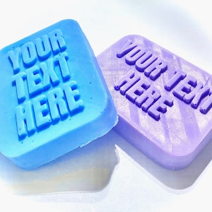 Puede incluir: Dos jabones cuadrados, uno azul claro y otro lavanda, con esquinas redondeadas. Cada jabón tiene el texto "YOUR TEXT HERE" en relieve en la superficie, en letras mayúsculas en negrita. Los jabones están sobre un fondo blanco.