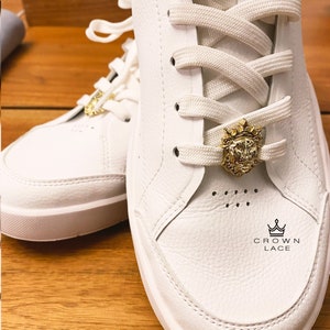 May include: White leather sneakers with white laces and a gold lion head charm on the laces. The charm has a crown on top. The text "CROWN LACE" is printed on the shoe.