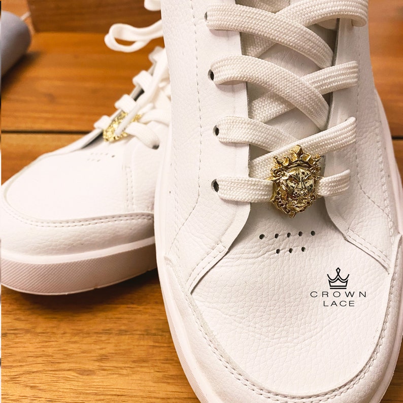 May include: White leather sneakers with white laces and a gold lion head charm on the laces. The charm has a crown on top. The text "CROWN LACE" is printed on the shoe.