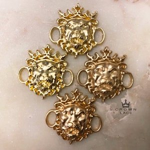 May include: Four gold-toned lion head shoe charms with a crown on top. Each charm has two loops for attaching to shoelaces. The charms are on a white surface with the text "CROWN LACE" in black.