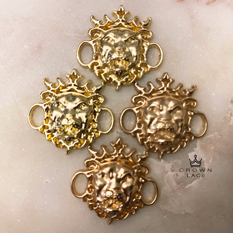 May include: Four gold-toned lion head shoe charms with a crown on top. Each charm has two loops for attaching to shoelaces. The charms are on a white surface with the text "CROWN LACE" in black.