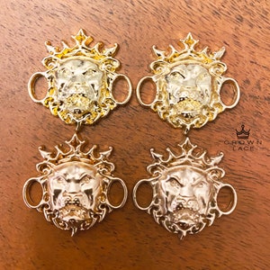 May include: Four gold-tone lion head shoe charms with a crown on top. Each charm has two loops for attaching to shoelaces. The charms are on a brown wooden surface.