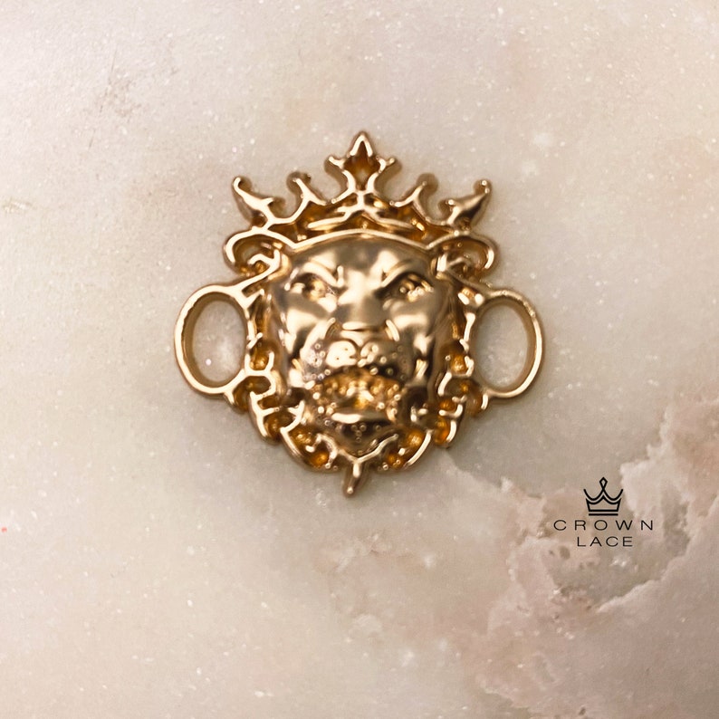 May include: Gold-toned lion head charm with a crown on top. The charm has two loops on the sides for attaching to a necklace or bracelet. The charm is on a white background.
