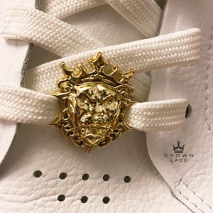 May include: A gold-colored lion head shoe lace charm with a crown on top. The charm is attached to a white shoelace. The text "CROWN LACE" is visible on the white leather shoe.