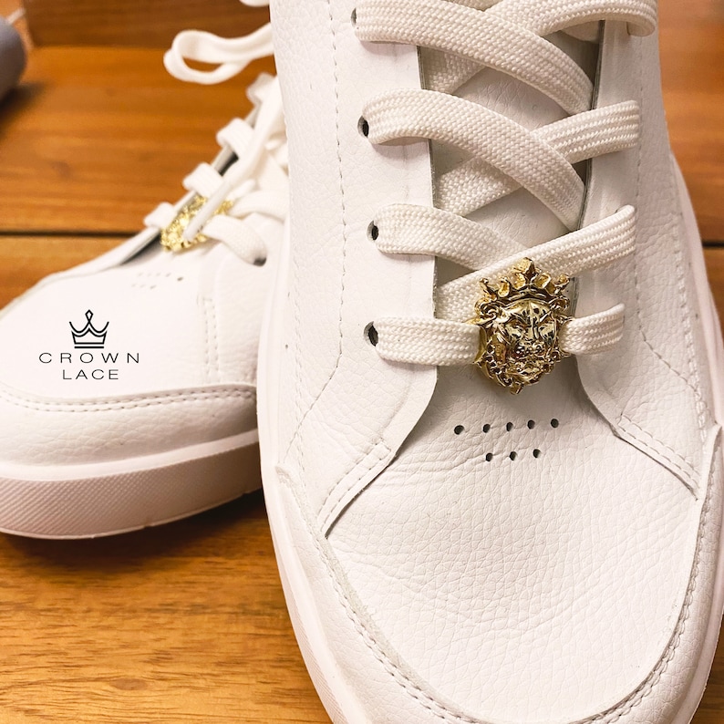May include: White leather sneakers with white laces and a gold lion head lace lock. The lace lock has a crown on top. The brand name "CROWN LACE" is printed on the side of the shoe.