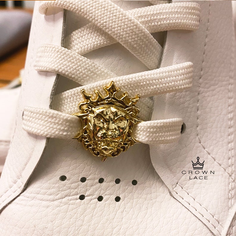 May include: White shoelaces with a gold lion head charm and a crown. The charm is attached to the laces with a loop. The text "CROWN LACE" is printed on the shoe.