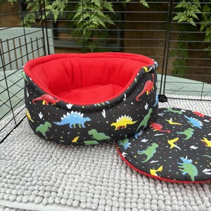 Peut inclure: Un lit pour animaux de compagnie noir avec un intérieur rouge et un coussin rond assorti. Le lit et le coussin sont décorés d'illustrations de dinosaures colorés. Le lit mesure environ 30 cm de diamètre.