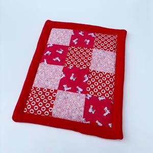 Peut inclure: Un tapis de jeu patchwork rouge et rose avec une bordure blanche. Le tapis est fait de différents tissus avec divers motifs, notamment de petits cercles blancs sur fond rouge, des motifs floraux roses et un motif de lamas blancs sur fond rose.