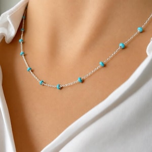 Op de afbeelding: Een delicate zilveren ketting met kleine, ronde turquoise kralen gelijkmatig verdeeld over een fijne ketting. De ketting wordt getoond tegen een wit overhemd, wat het levendige blauw van de kralen benadrukt.