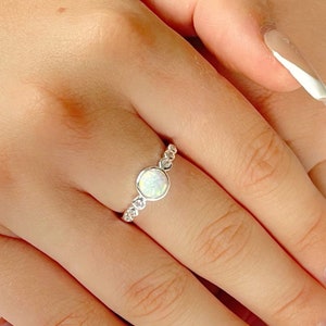 May include: A silver ring featuring a white opal stone and small clear stones around it. The ring is worn on a finger with white and floral nail polish.