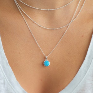 May include: A layered silver necklace with a turquoise stone pendant. The necklace is worn by a person with long brown hair.