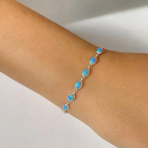 May include: A silver chain bracelet featuring a series of seven oval-shaped, iridescent blue opal stones set in individual metal bezels, connected by small circular silver links, creating a delicate and continuous line of blue gemstones.