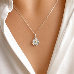 May include: A silver daisy pendant necklace with a yellow center. The pendant is suspended from a delicate silver chain. The necklace is worn against a white shirt.