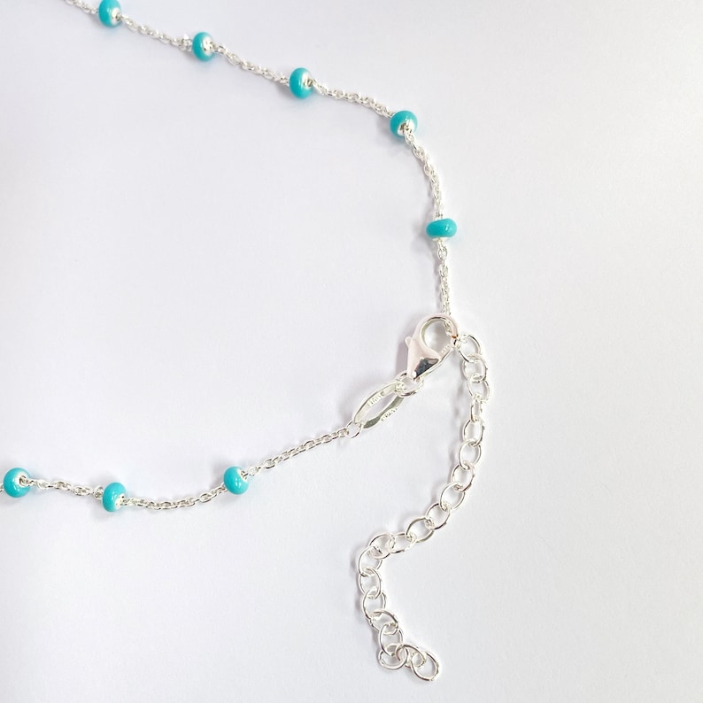 Op de afbeelding: Een zilveren ketting met kleine, turquoise kralen gelijkmatig verdeeld over de ketting. De ketting heeft een karabijnsluiting en een verstelbare ketting. De achtergrond is effen wit. Geschikt voor diverse gelegenheden.