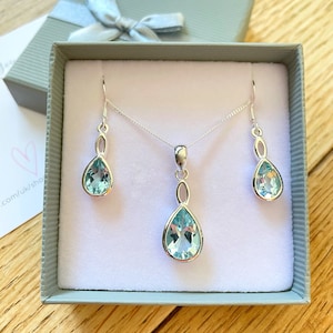 May include: A silver necklace and earrings set featuring teardrop-shaped blue topaz stones. The necklace has a delicate chain and a pendant with a twisted design. The earrings are drop earrings with a similar twisted design.