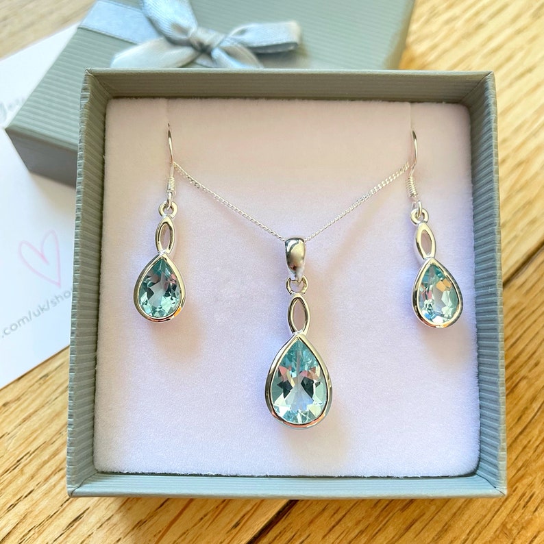 May include: A silver necklace and earrings set featuring teardrop-shaped blue topaz stones. The necklace has a delicate chain and a pendant with a twisted design. The earrings are drop earrings with a similar twisted design.