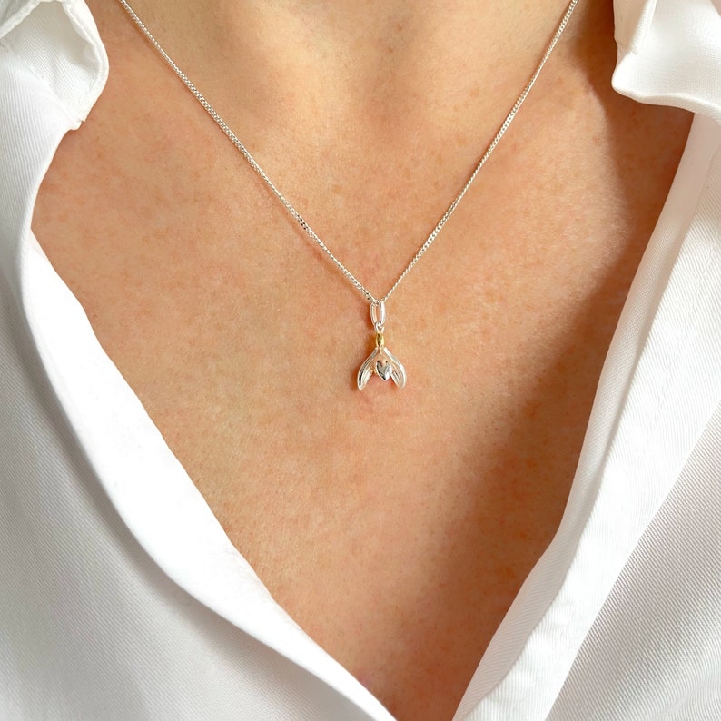 Op de afbeelding: Een zilveren ketting met een delicate hanger in de vorm van een sneeuwklokje. De hanger is gemaakt van zilver en goud, met een klein hartje in het midden.