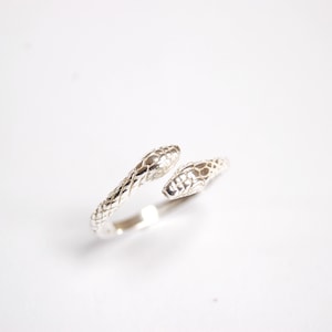 May include: A silver ring with two snake heads facing opposite directions. The ring is made of a smooth, shiny metal and has a delicate design.