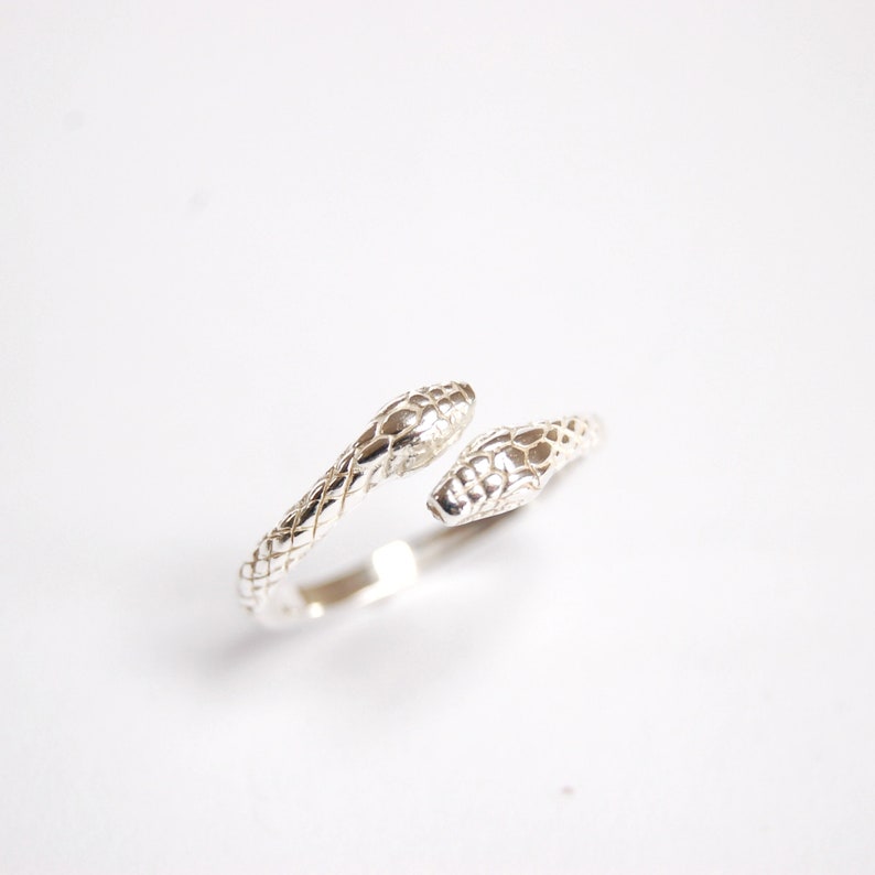 May include: A silver ring with two snake heads facing opposite directions. The ring is made of a smooth, shiny metal and has a delicate design.