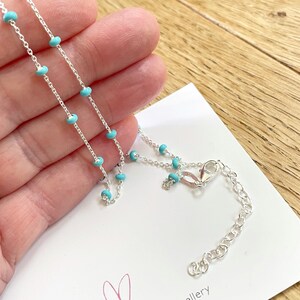 Op de afbeelding: Een delicate zilveren ketting met kleine, turquoise kralen gelijkmatig verdeeld over de ketting. De ketting heeft een karabijnsluiting en een verlengketting. De sieraden worden gepresenteerd op een witte kaart.
