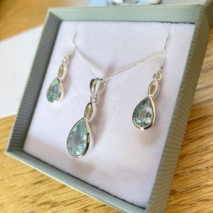 May include: A silver necklace and earrings set with teardrop-shaped blue gemstones. The jewellery is displayed in a white box.
