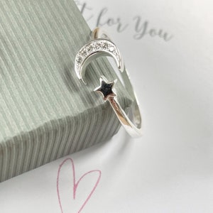 May include: Silver ring featuring a crescent moon design with a star. The moon is decorated with small clear stones.