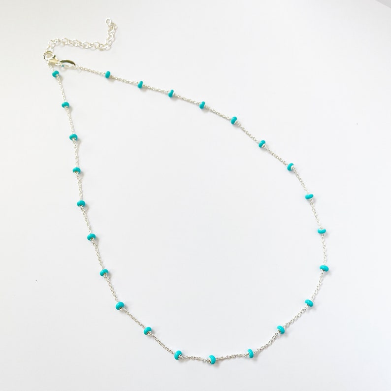 Op de afbeelding: Een delicate zilveren ketting met kleine, ronde turquoise kralen verdeeld over een fijne ketting. De ketting heeft een kreeftsluiting en wordt getoond tegen een witte achtergrond. Dit sieraad is geschikt voor dagelijks gebruik.