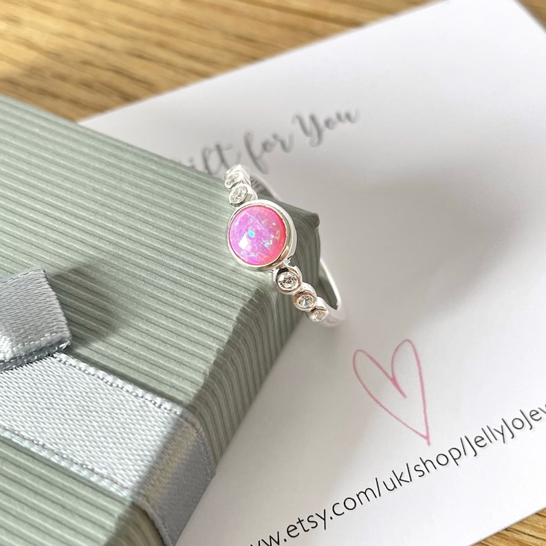 Op de afbeelding: Een zilveren ring met een roze opaal-edelsteen en een band van kleine, heldere stenen. De ring zit in een cadeaudoos met een label waarop staat "Cadeau voor jou".