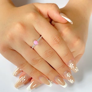 May include: A silver ring with a pink opal gemstone. The ring is on a finger with a white french manicure and daisy nail art.