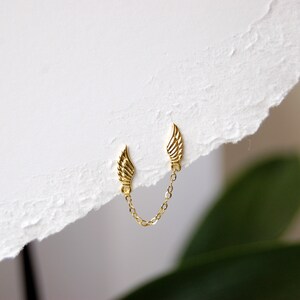 May include: A pair of gold wing earrings connected by a delicate gold chain. The earrings are small and delicate, and the chain is about 1 inch long.