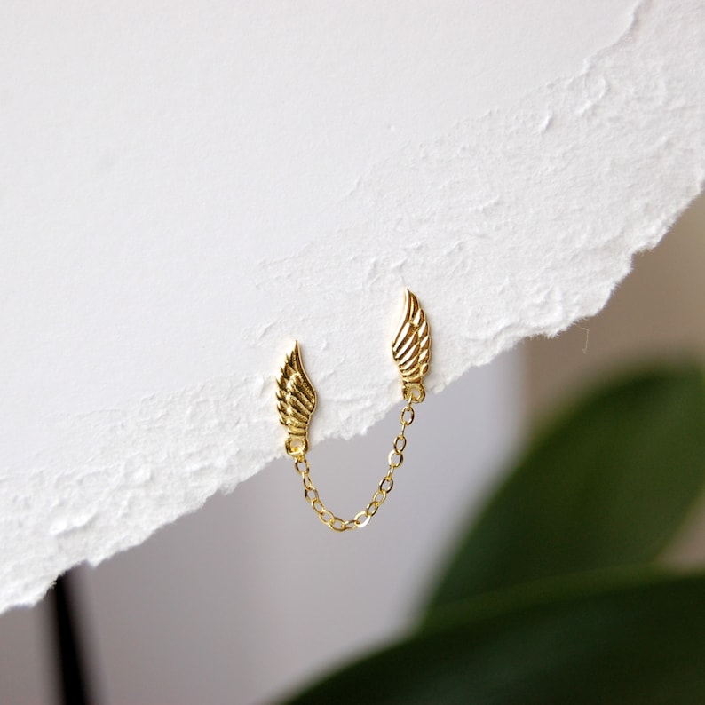 May include: A pair of gold wing earrings connected by a delicate gold chain. The earrings are small and delicate, and the chain is about 1 inch long.