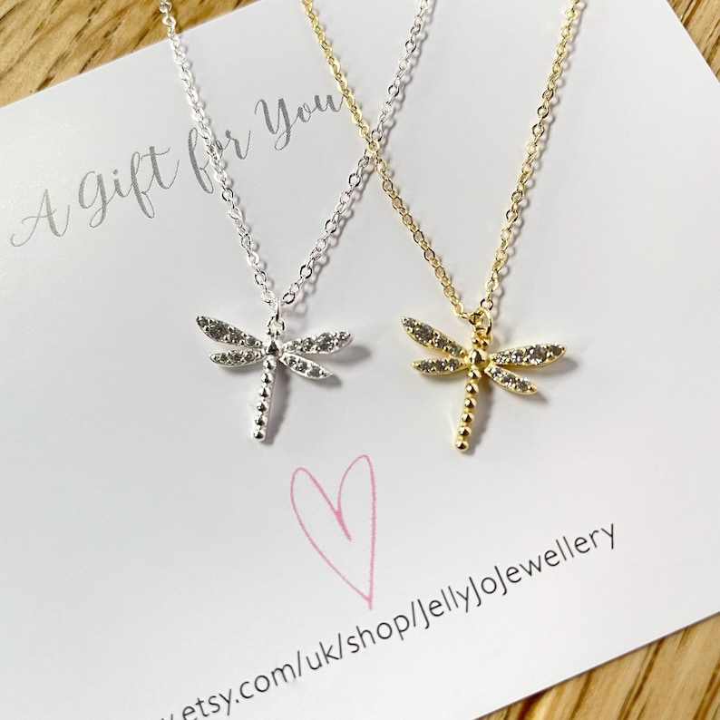 May include: Two dragonfly necklaces, one silver-toned and one gold-toned, each featuring clear stones on the wings and a beaded body. The necklaces are displayed on a white card with the text A Gift for You and a pink heart illustration.