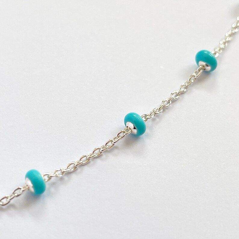 Op de afbeelding: Een delicate zilveren ketting met drie turquoise kralen gelijkmatig verdeeld over de lengte. De kralen hebben een levendige, opvallende kleur en de ketting heeft een fijn, gedetailleerd schakelpatroon.