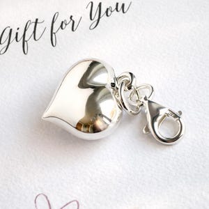 May include: A shiny silver heart-shaped charm with a lobster clasp and connecting rings. The charm is reflective and rests on a white surface with the words "gift for you" in cursive script.