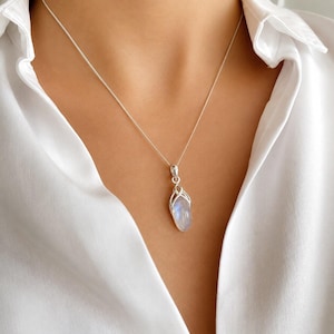 May include: A silver necklace with a pendant featuring a teardrop-shaped, iridescent gemstone. The pendant is encased in a decorative silver setting and hangs from a delicate silver chain. The necklace is displayed against a white shirt.