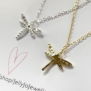 May include: Two dragonfly necklaces, one silver-toned and one gold-toned, each featuring a delicate chain and a pendant with small embedded stones. A hand-drawn pink heart and the text shop/jellyjojewellery are visible on the white surface.