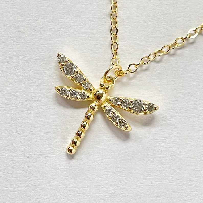May include: A gold-toned necklace featuring a dragonfly pendant. The dragonfly has a segmented body and four wings encrusted with small, clear, sparkling stones. The pendant is attached to a delicate gold-toned chain with small, round, open links.