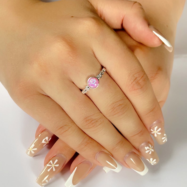 Op de afbeelding: Een zilveren ring met een roze opaal-steen en kleine heldere stenen rond de band. De ring wordt gedragen op een vinger met lichtroze nagellak en witte tips.