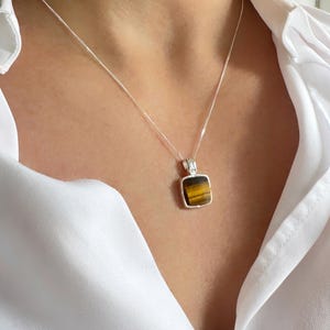 May include: A silver necklace with a square pendant featuring a tiger's eye stone. The pendant is set in a silver frame and hangs from a delicate silver chain. The tiger's eye stone displays bands of brown and gold.