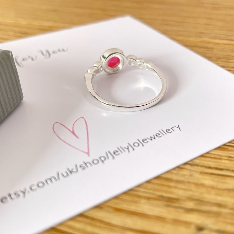 Op de afbeelding: Een zilveren ring met een roze edelsteen in een bezel-setting. De ring heeft een eenvoudige band en is versierd met kleine heldere stenen aan weerszijden van de edelsteen.