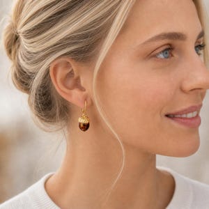Könnte beinhalten: Goldfarbene Eichel-Ohrringe mit einem braunen, gesprenkelten Körper und einer strukturierten Kappe. Die Ohrringe hängen an einem einfachen Bügel. Die Ohrringe sind ein Accessoire zum Thema Herbst.
