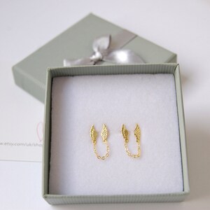 May include: A pair of gold-tone earrings with a delicate chain connecting two small wing charms. The earrings are displayed in a white box with a gray lid.